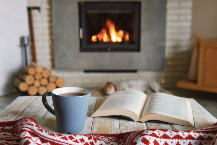 mug and open book set on a table in front of lit fireplace with a stack of wood next to it