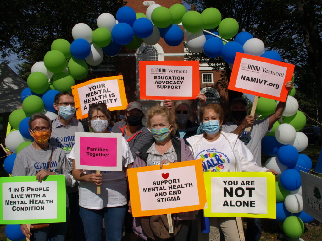 eight adults wearing masks stand under an archway of green blue and white balloons holding signs in support of mental health