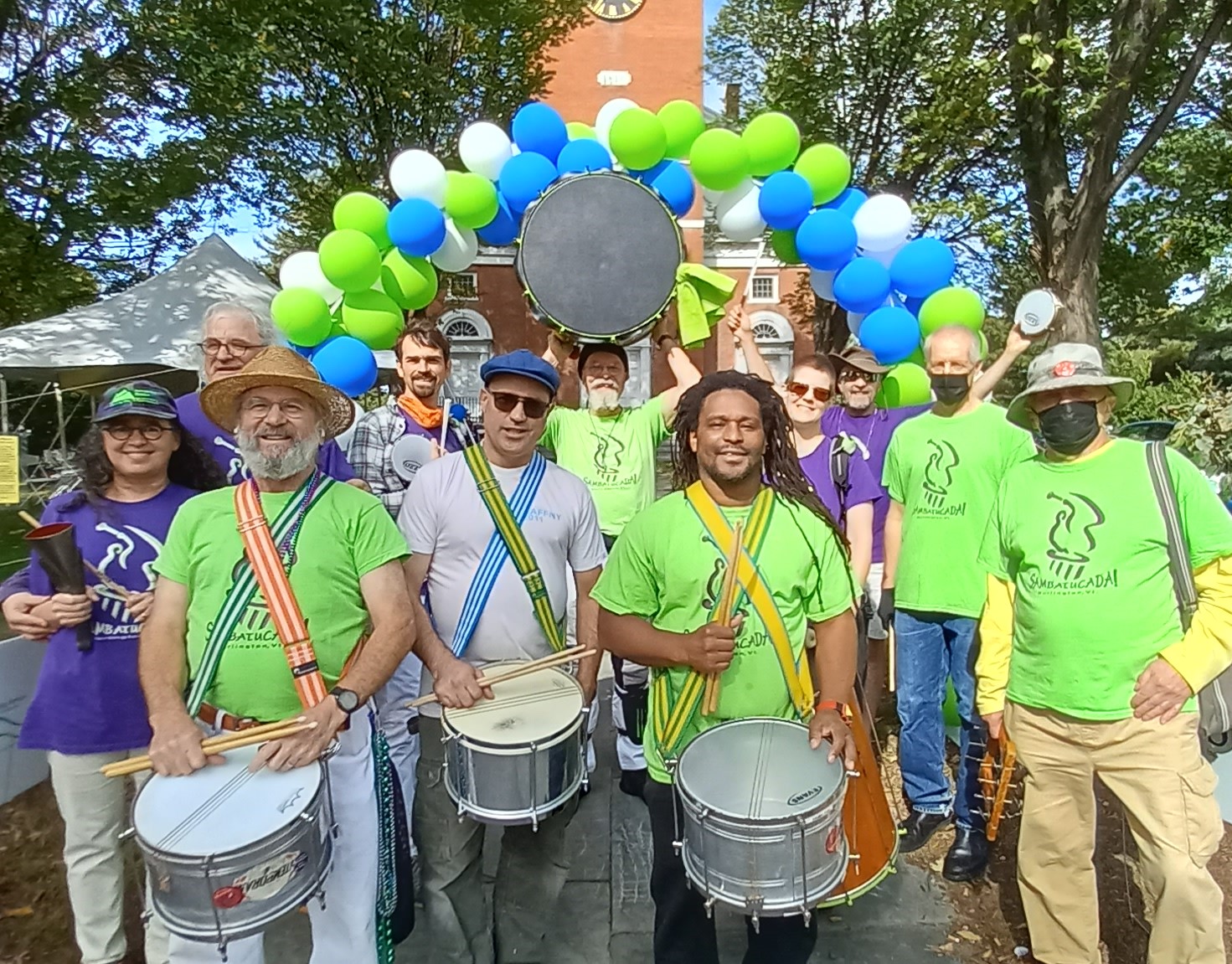 group of adults with drums stand in front of a green and blue balloon arch